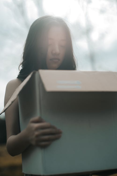 Young dreamy female with black hair and closed eyes carrying carton box in sunlight