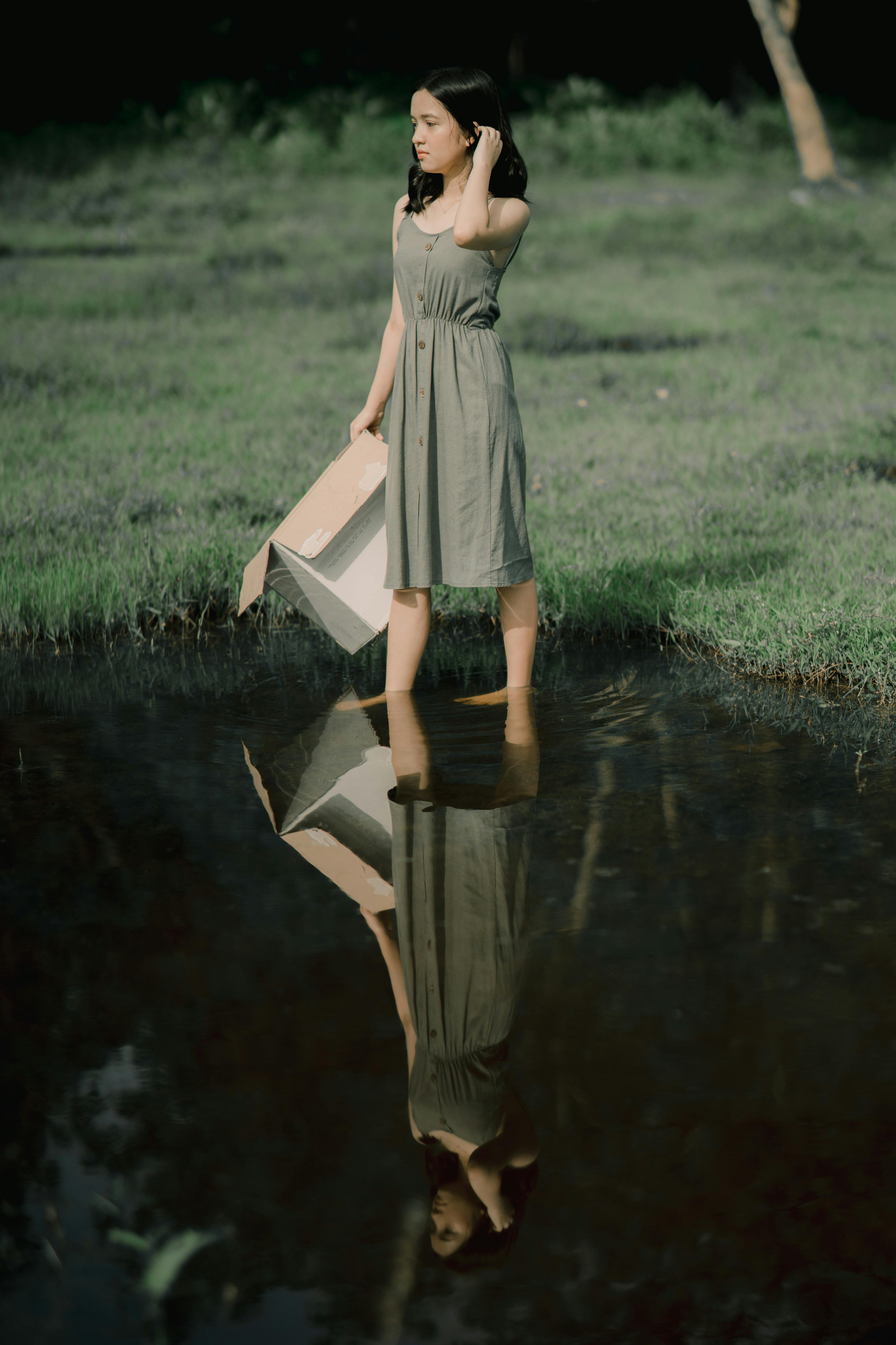 Young contemplative female in sundress with cardboard box touching hair while reflecting in water and looking away
