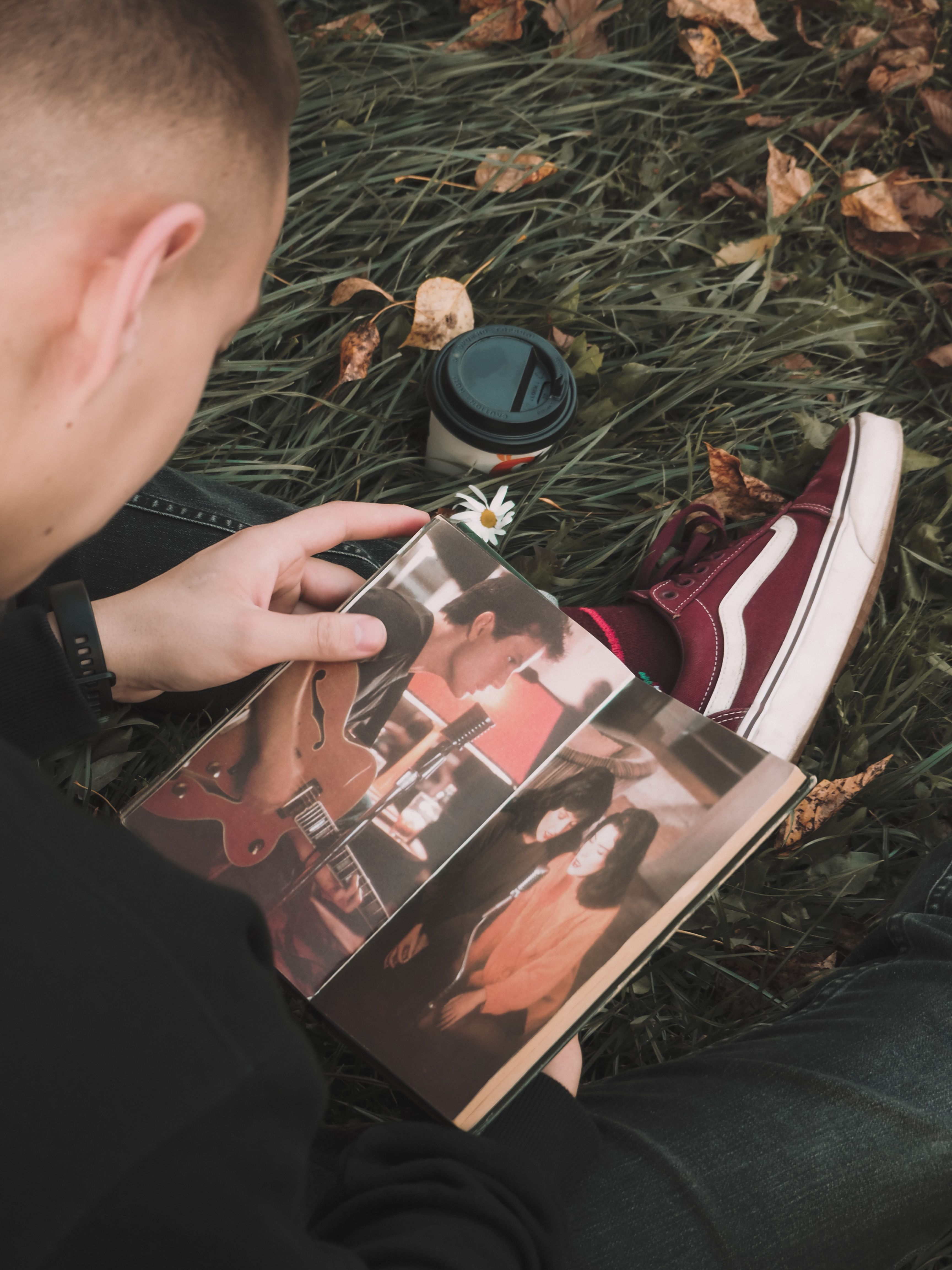 A young man sits on grass in autumn, enjoying reading with a coffee nearby.