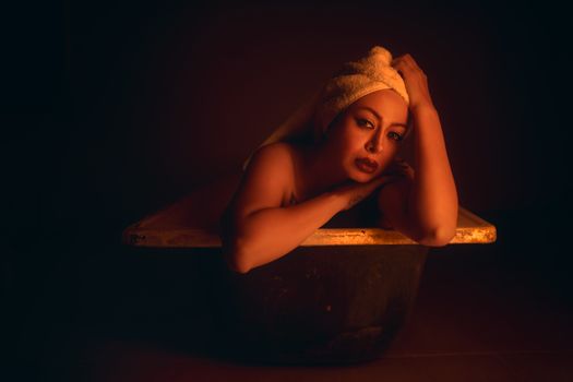 A dramatic portrait of a woman in a bathtub with a towel wrapped around her head under low light conditions.