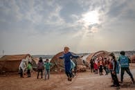 Full body of many poor children walking and talking near tents under sunny cloudy sky