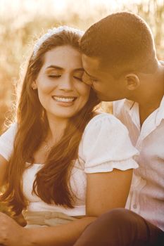 Gentle ethnic boyfriend kissing and caressing smiling girlfriend sitting in nature together in sunny romantic day