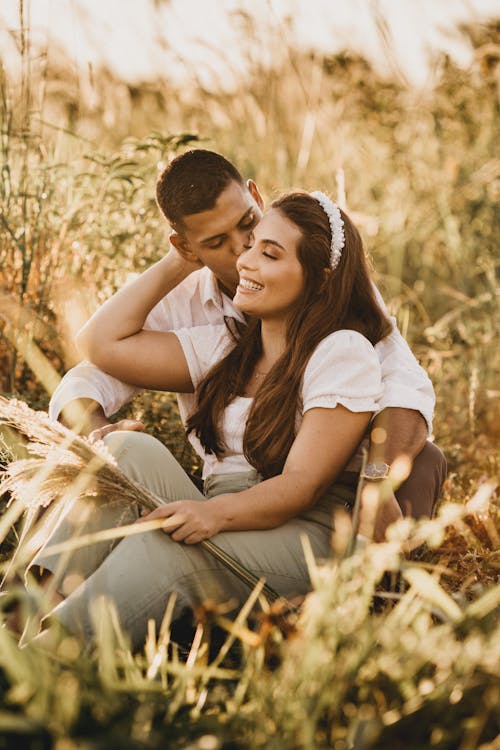 Free Loving diverse couple hugging and kissing sitting close together in grassy meadow in sunlight and enjoying romantic moment Stock Photo