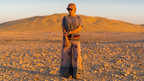 Full body of calm young female traveler in casual clothes and sunglasses standing on barren terrain during trip in desert at sundown