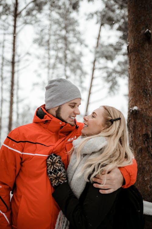 Smiling couple in warm outerwear hugging gently and happily smiling at each other in snowy woods