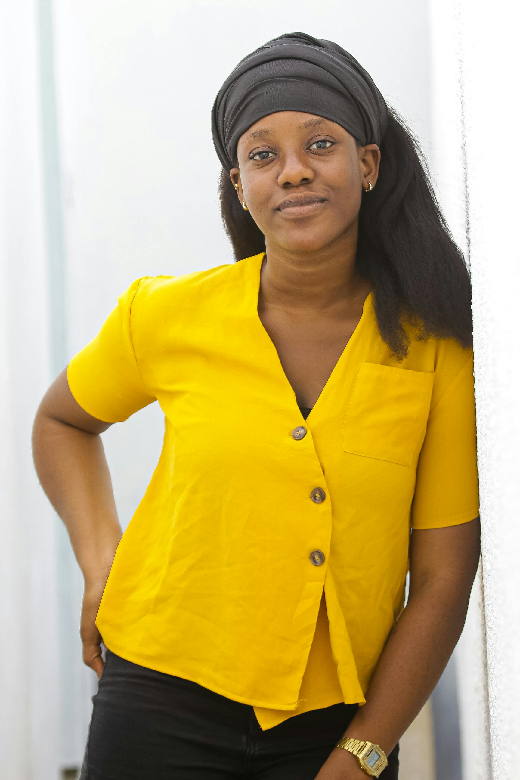 Portrait of a smiling woman in a yellow top and head scarf leaning against a wall indoors.