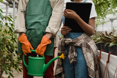 Two people in a greenhouse with a watering can and clipboard