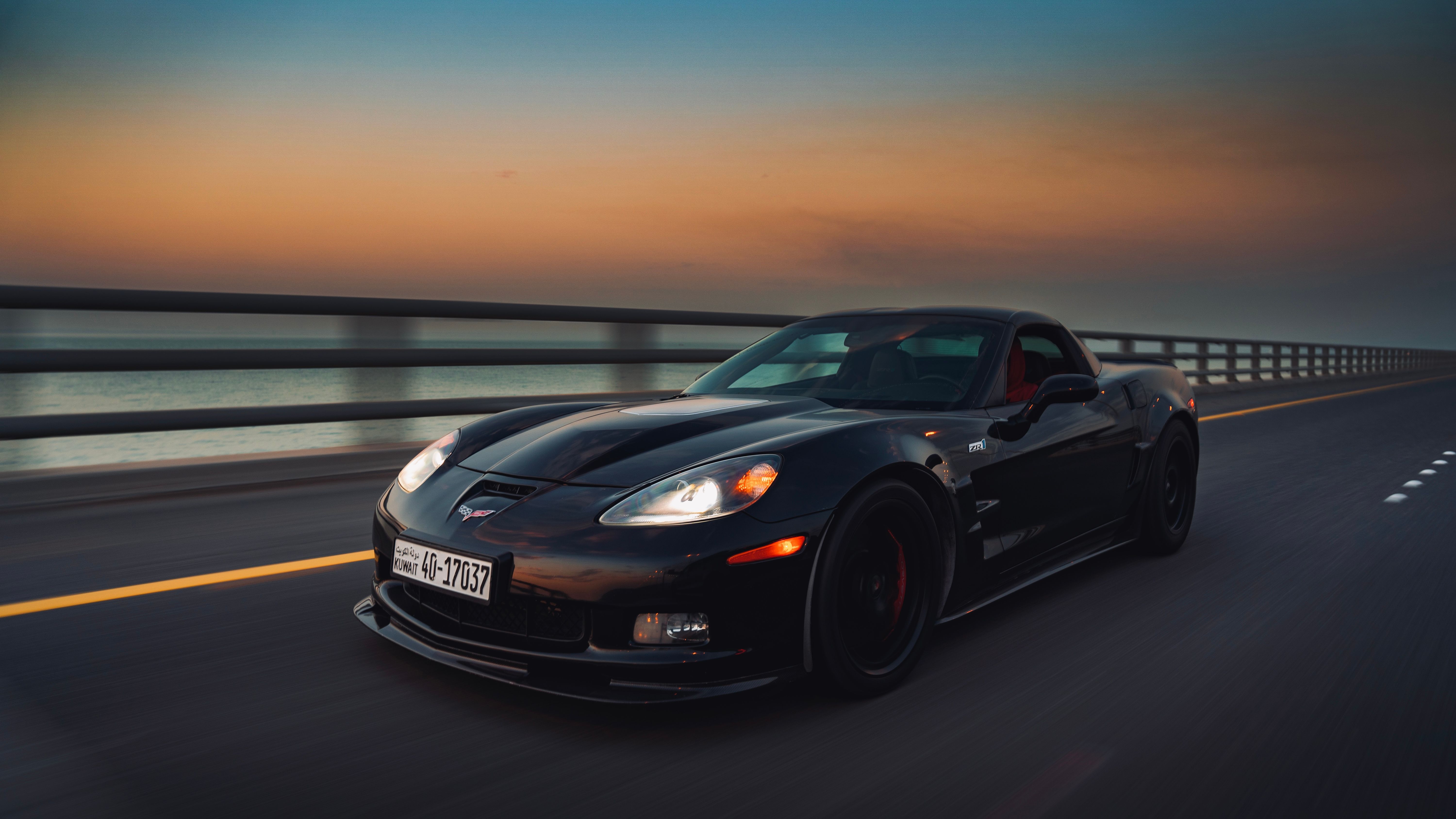 A sleek black sports car speeds along a coastal bridge at twilight, captured in motion.