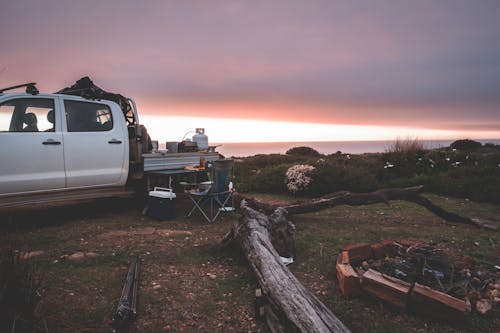 Modern pickup truck parked on glade near unfolded travel chair table and round bonfire place in peaceful vast nature under amazing evening sky