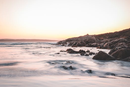A tranquil sunset scene over a rocky coastline with calm ocean waves and a pastel sky.