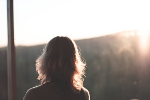 Back view of unrecognizable female traveler with blond hair standing near window and admiring mountains on sunny day