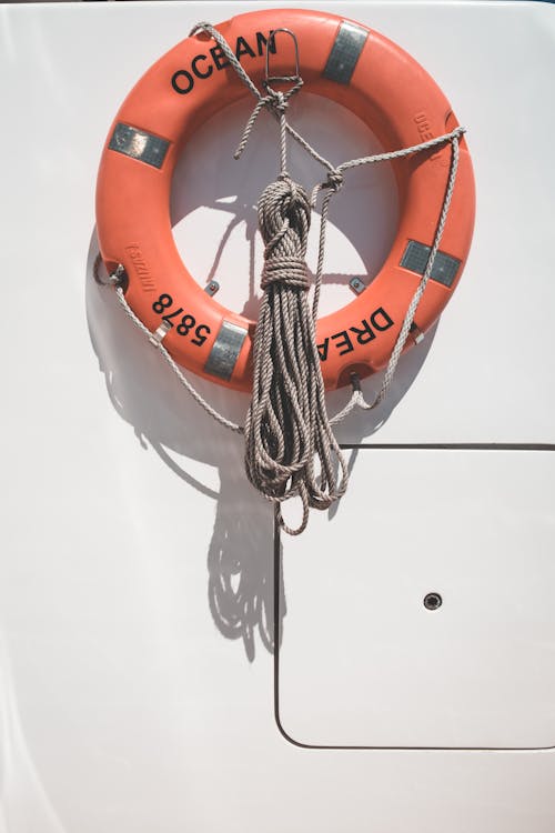 Orange lifebuoy with tied mooring rope hanging on white wall of modern yacht on sunny day