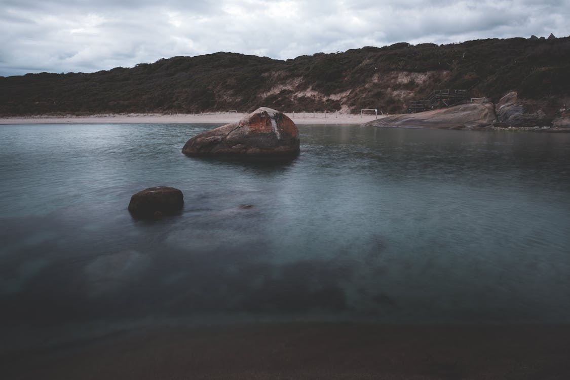Free Picturesque landscape of rocky boulders in calm ocean water near sandy beach surrounded by hills covered with lush green trees against cloudy sky Stock Photo
