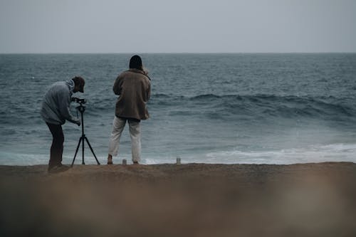 Unrecognizable men photographers in casual clothes standing on beach and taking photo on professional photo camera of stormy sea in daylight