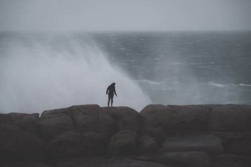 Back view of unrecognizable male standing on rocky cliff and admiring stormy sea with splashes in cloudy day