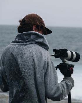 Back view of photographer in casual clothes and cap standing with tripod with professional photo camera and shooting wavy sea in cloudy day