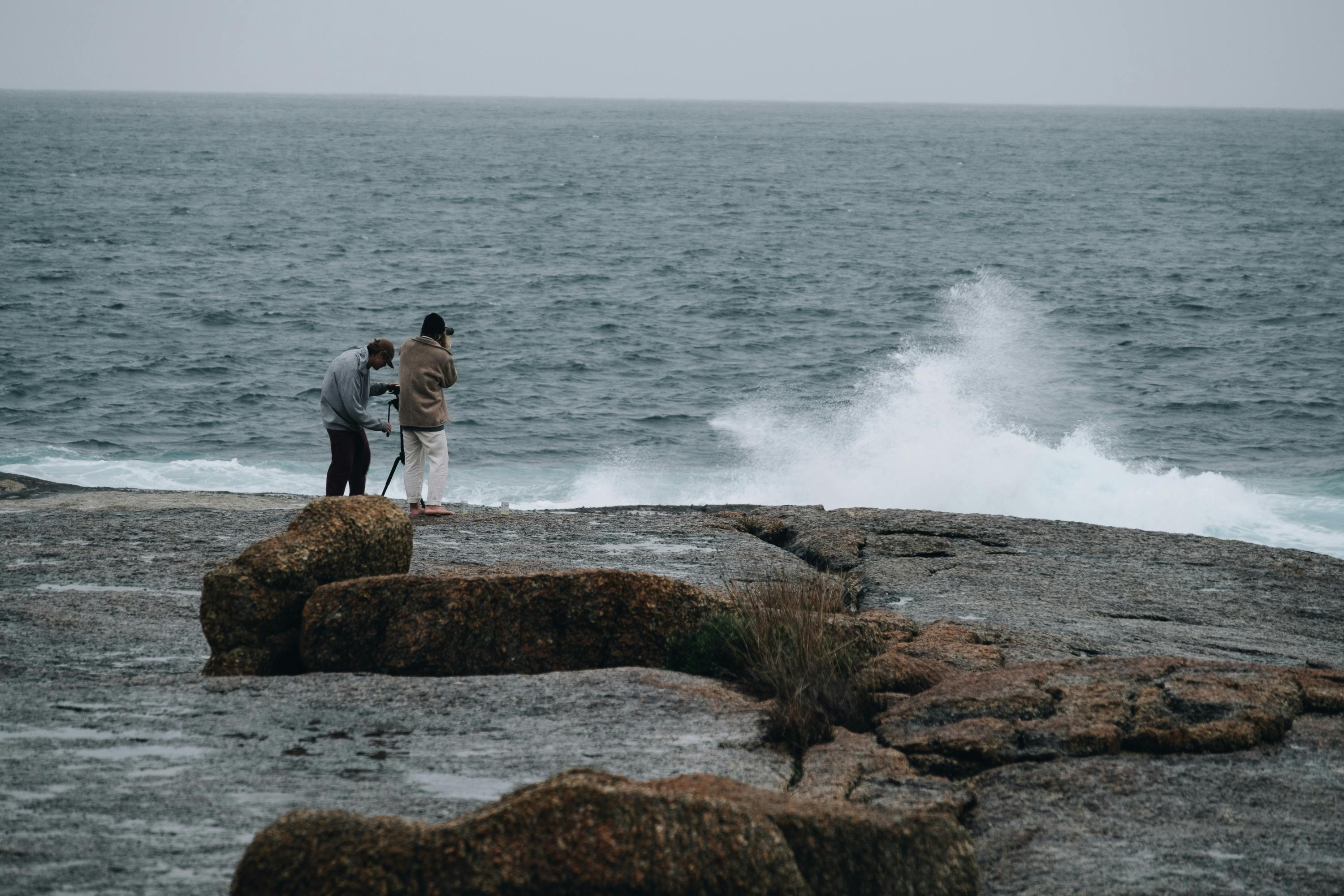 Side view of anonymous photographers standing on wet rocky shore and taking photos of splashing waves of ocean in cloudy day
