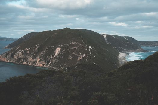 Breathtaking scenery of Isthmus Hill and Bald Head walking track covered with lush green vegetation and washing by foamy ocean against cloudy sky in Albany