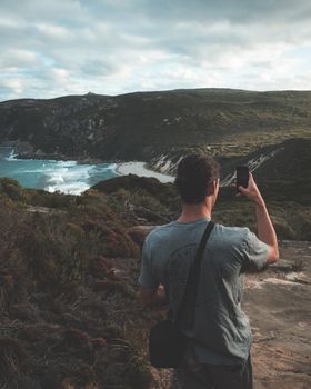 Back view of unrecognizable male traveler in casual clothes using smartphone while taking photo of picturesque foamy ocean surrounded by green hills during trekking against overcast sky