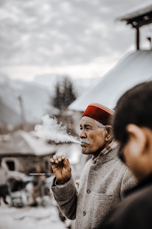 Thoughtful elderly Asian male in red traditional hat smoking cigarette and looking away while standing on street in snowy winter village