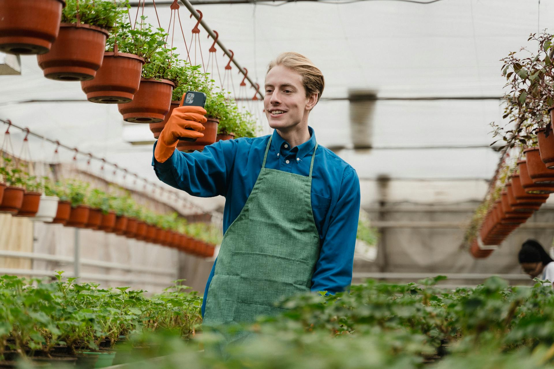 Smiling gardener taking a selfie in a greenhouse filled with potted plants.