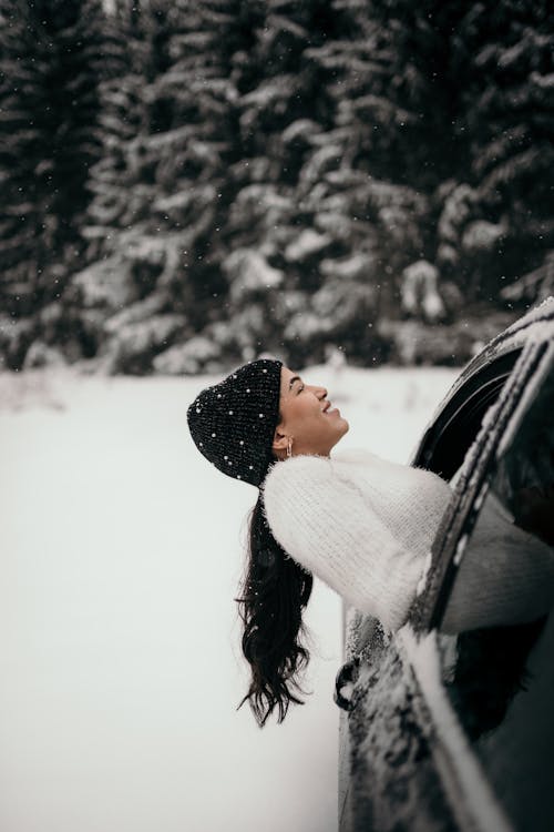 Side view of cheerful female with long hair wearing warm hat and sweater enjoying snowfall from car window in woods