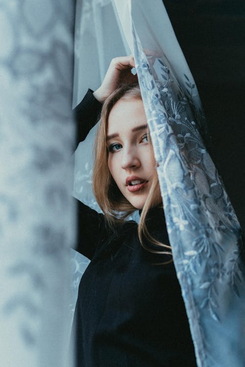 Low angle of sensual young female standing behind curtain and looking at camera