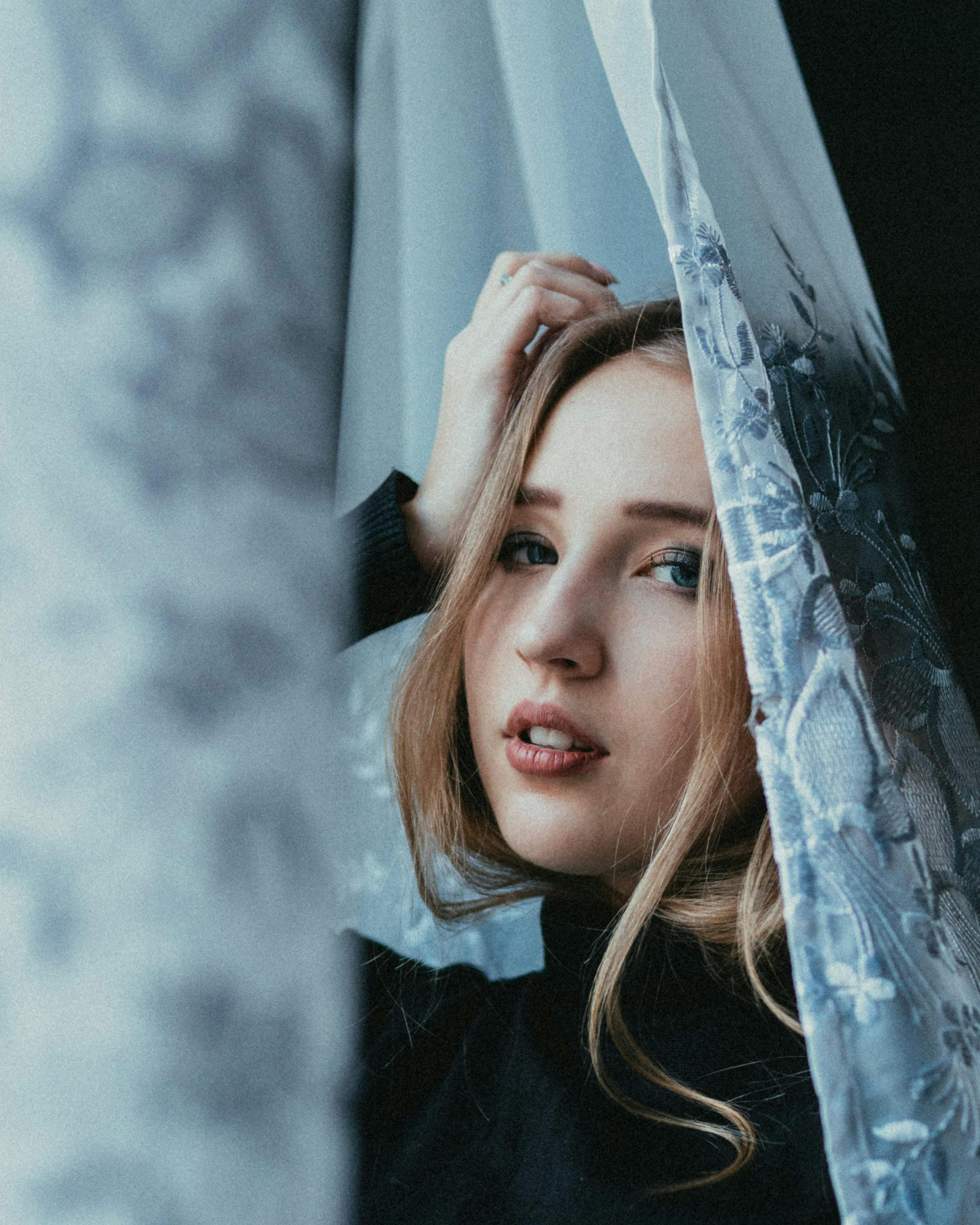 Charming young female leaning on hand standing near curtains and looking at camera