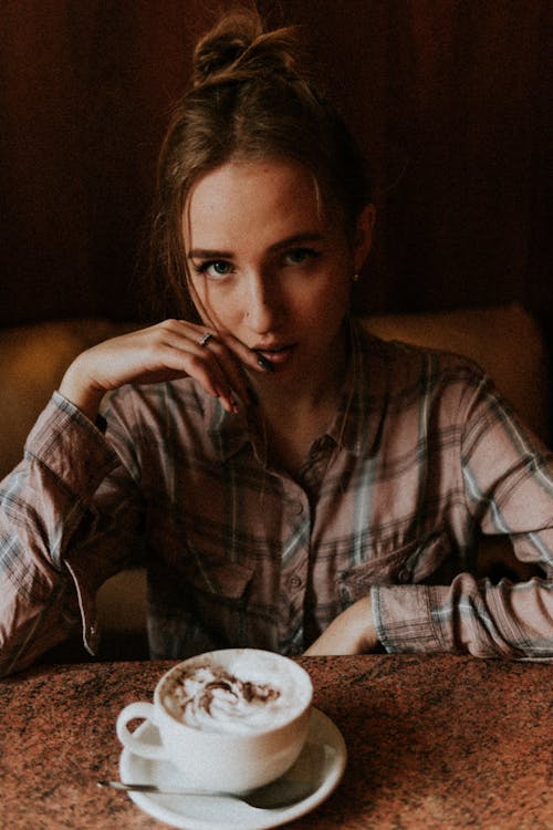 High angle of serious female touching face sitting at table with cappuccino with froth in coffee shop