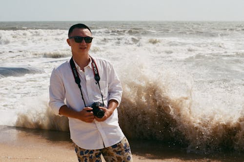 Free Young Man with a Camera Standing on a Beach with Big Waves Behind Him  Stock Photo