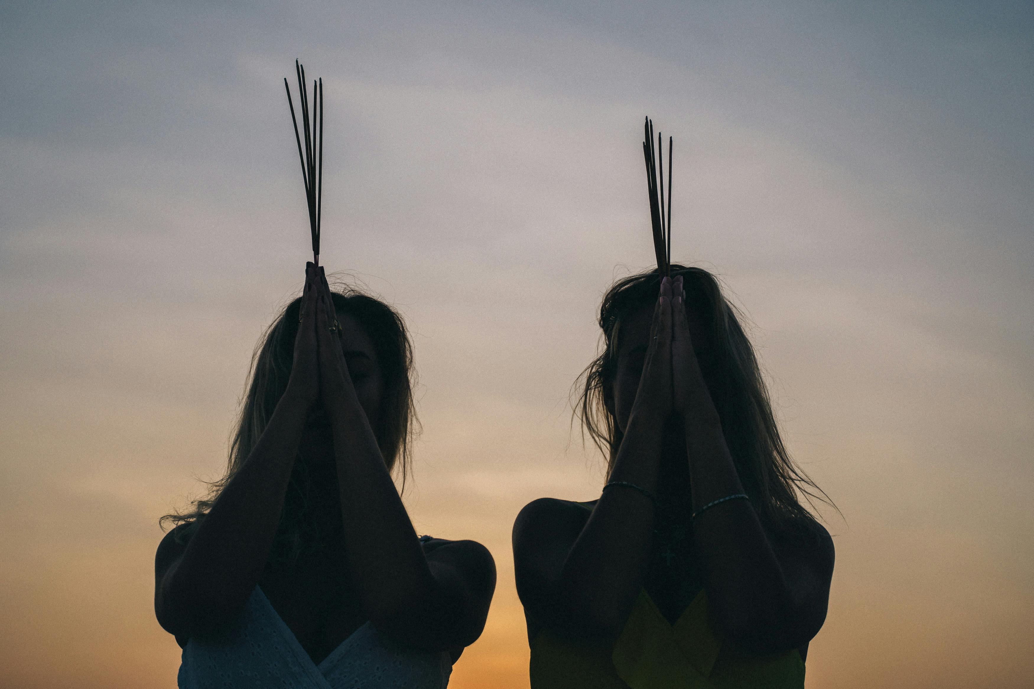 Two women meditating at sunset with incense sticks, creating a calming silhouette scene.