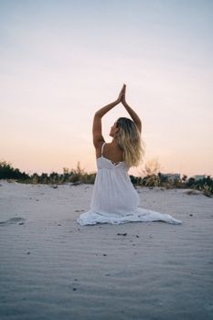A woman in a white dress practicing yoga on a sandy beach at sunset, creating a tranquil scene.