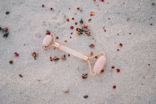 Rose quartz facial roller on sandy beach with dried petals, promoting relaxation and skincare.