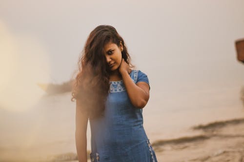 Content Indian female with long hair in dress looking at camera while standing on sandy beach near sea with wooden boat
