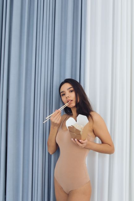 A woman in a swimsuit eating Asian noodles with chopsticks in a stylish indoor setting.