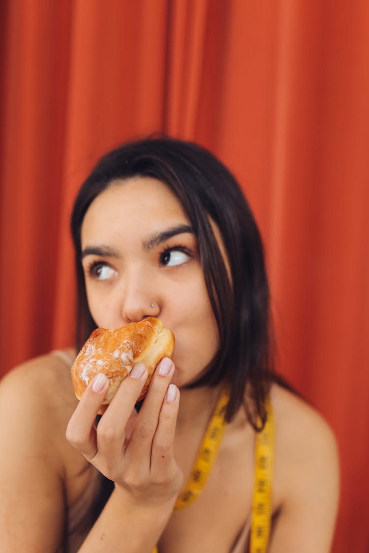 A Woman With Tape Measure Wrapped Around Her Neck Eating Donut While Looking Afar