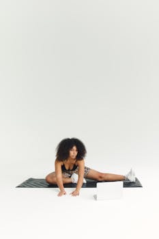 African American woman stretching on a yoga mat using a laptop indoors.