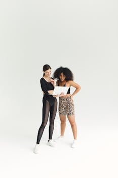 Two women in activewear using a laptop, smiling and standing in a studio.