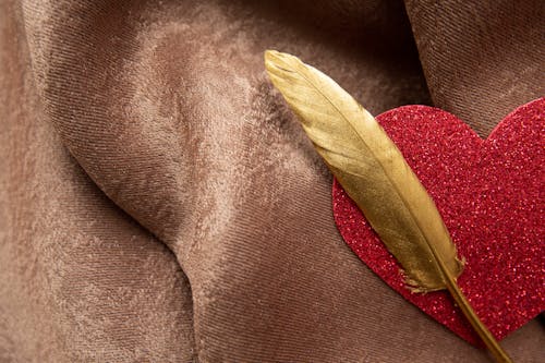 Top view of shiny decorative red heart with long golden feather placed on soft brown plaid during festive event celebration