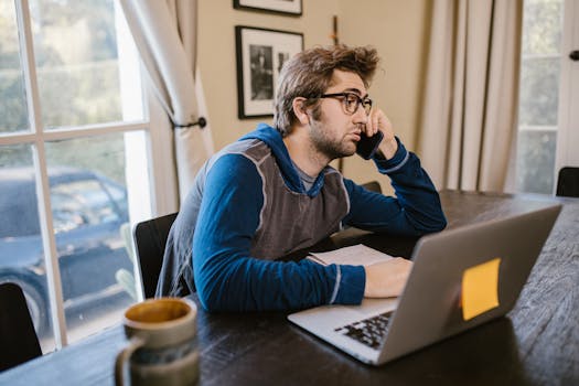A young man with glasses working from home on his laptop, looking tired and thoughtful.