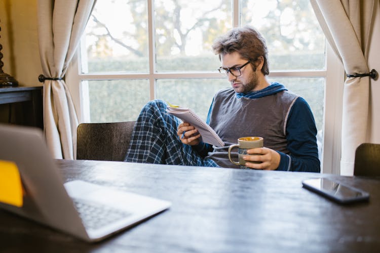 A Man Sitting Near The Table While Holding A Notebook And A Cup Of Coffee