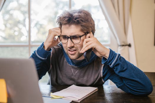 Man in glasses using laptop and smartphone at home, appearing confused.