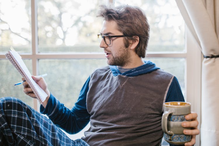 Man Holding A Ceramic Mug While Reading A Notebook 