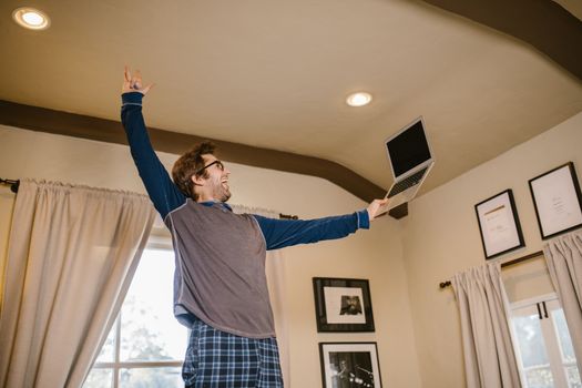 An excited man joyfully raises his laptop while standing in a cozy room.