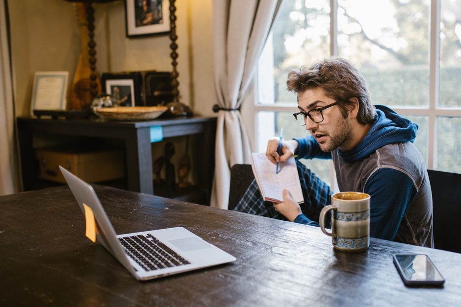 A young man in casual attire taking notes while working from home with a laptop.
