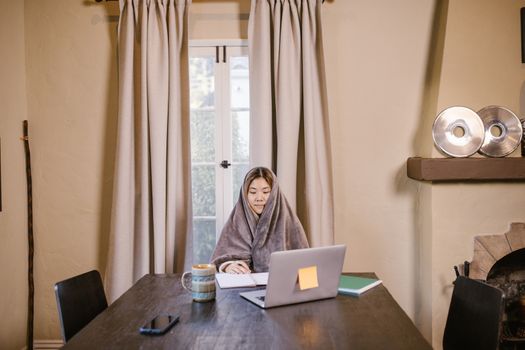 Asian woman cocooned in a blanket, working on a laptop with coffee at home office.