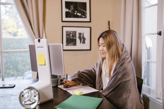 Asian woman focused on online learning at home, wrapped in a blanket with a notebook and computer.