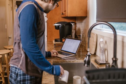 Adult man multitasking at kitchen with a laptop and a cup, blending work and home life.