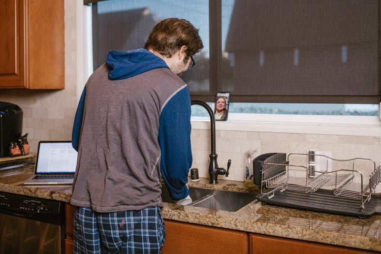 A Man Standing Near The Kitchen Sink While Doing Video Call On His Mobile Phone