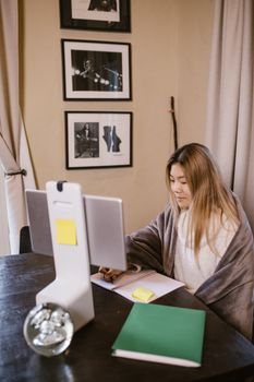 Asian woman working remotely, wrapped in a gray blanket, indoors at a desk. Cozy home office environment.